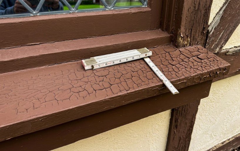 Brown paint on a wooden window sill cracked into a deep scaly alligator pattern, with a folding ruler for scale