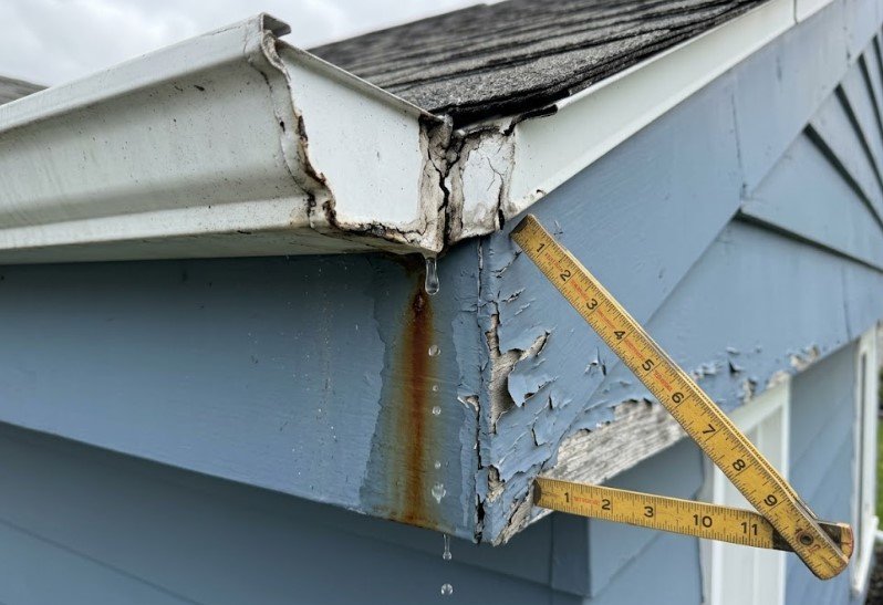Split gutter corner seam dripping water onto peeling blue trim, with a folding ruler for scale