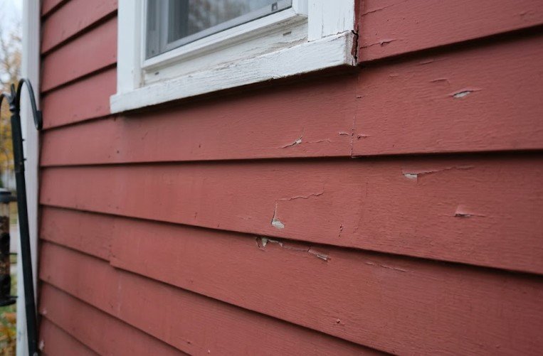 Flaking red paint on wood lap siding below a window, with a split joint at the corner of the sill
