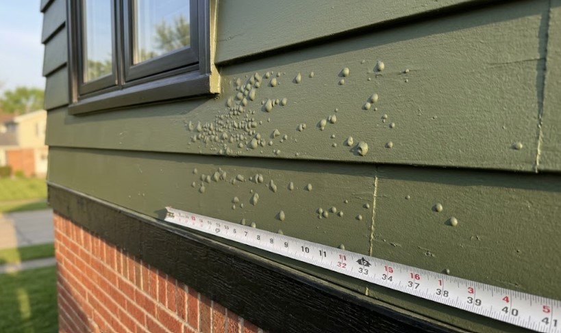 Clusters of paint blisters on lap siding below a window, with a tape measure for scale