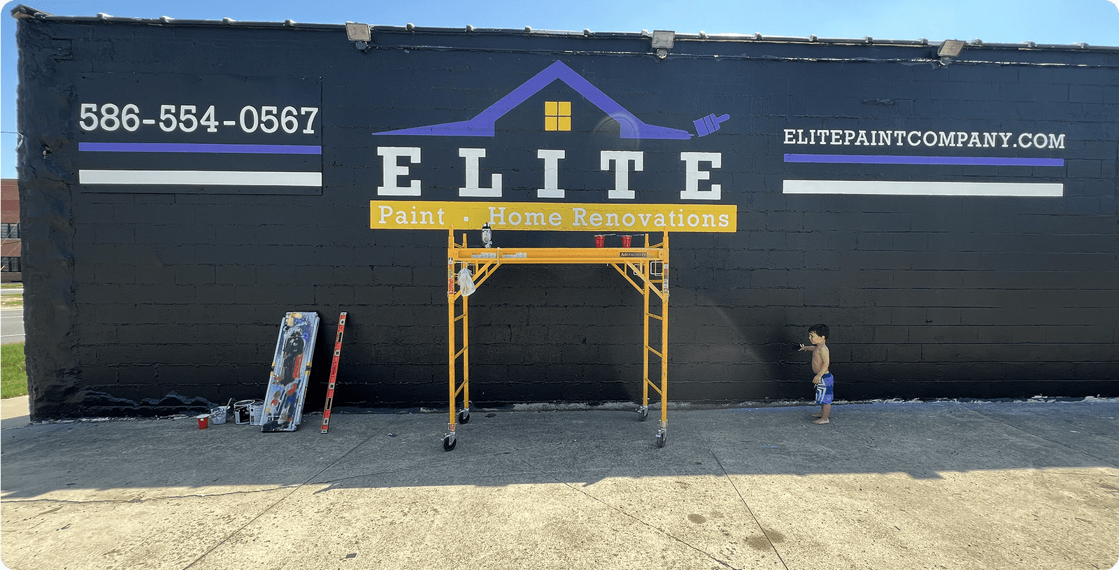 Black wall with "Elite Paint Home Renovations" ad, scaffold, painting tools, and a child nearby.