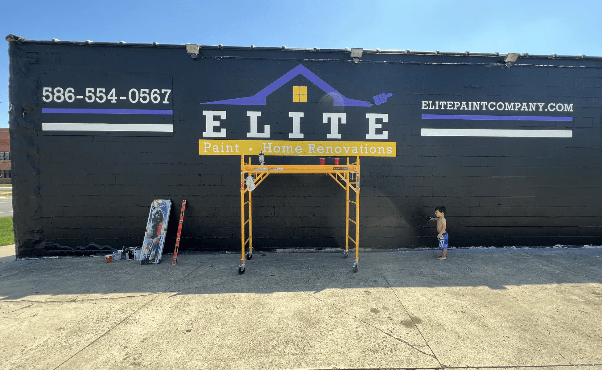 Painted wall with Elite Paint and Home Renovations logo, scaffold, tools, and child nearby.
