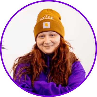 Person with long reddish-brown hair, mustard beanie, purple sweater, on plain background, purple border.