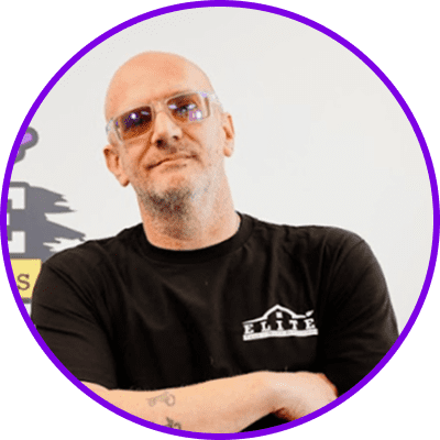 Home 25 Person with shaved head and glasses in a black "Elite" t-shirt, arms crossed, tattoo visible.