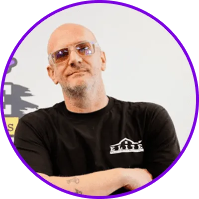 Person with shaved head and glasses in a black "Elite" t-shirt, arms crossed, tattoo visible.