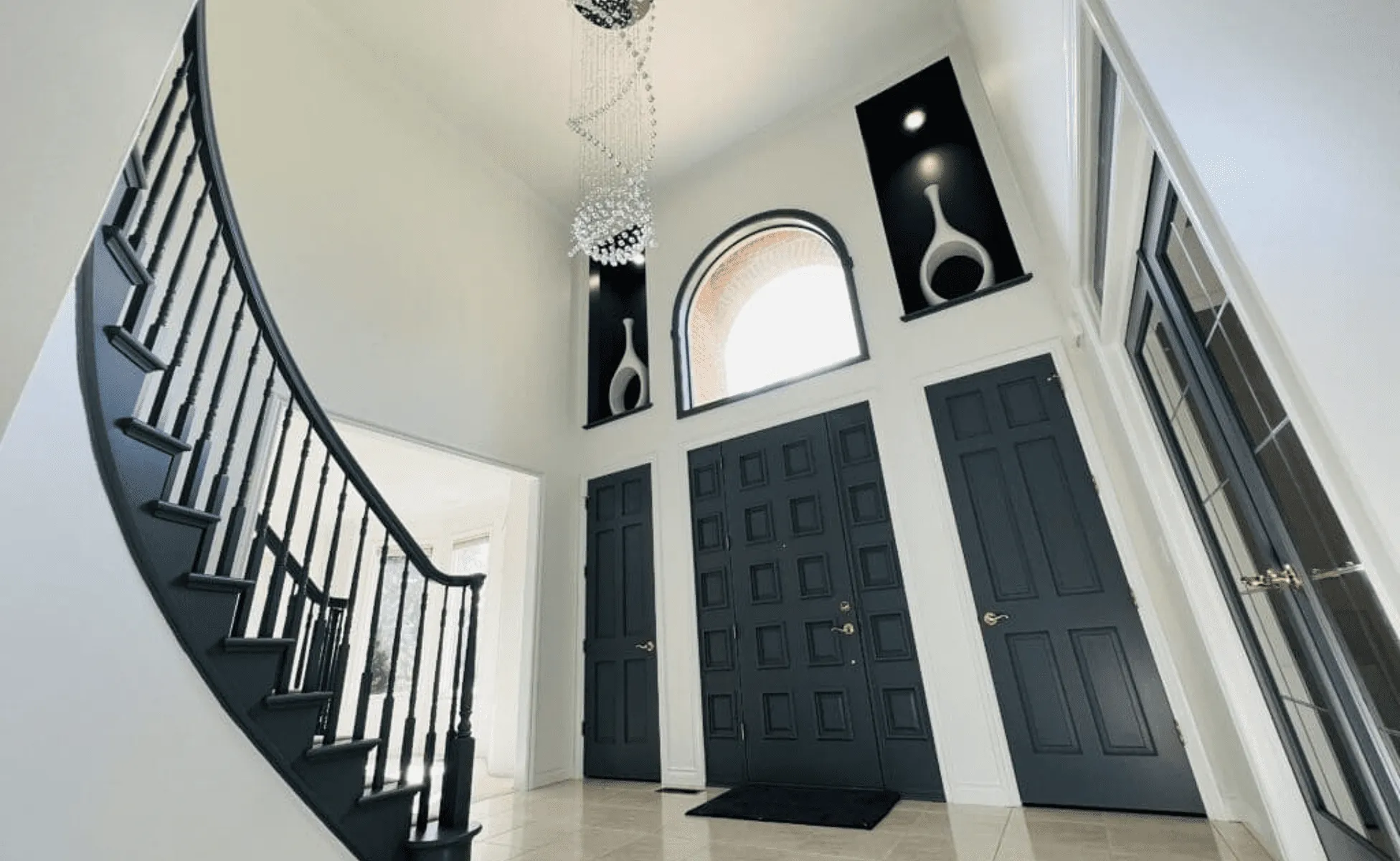 Spacious foyer with dark staircase, double doors, chandelier, art niches, and arched window.