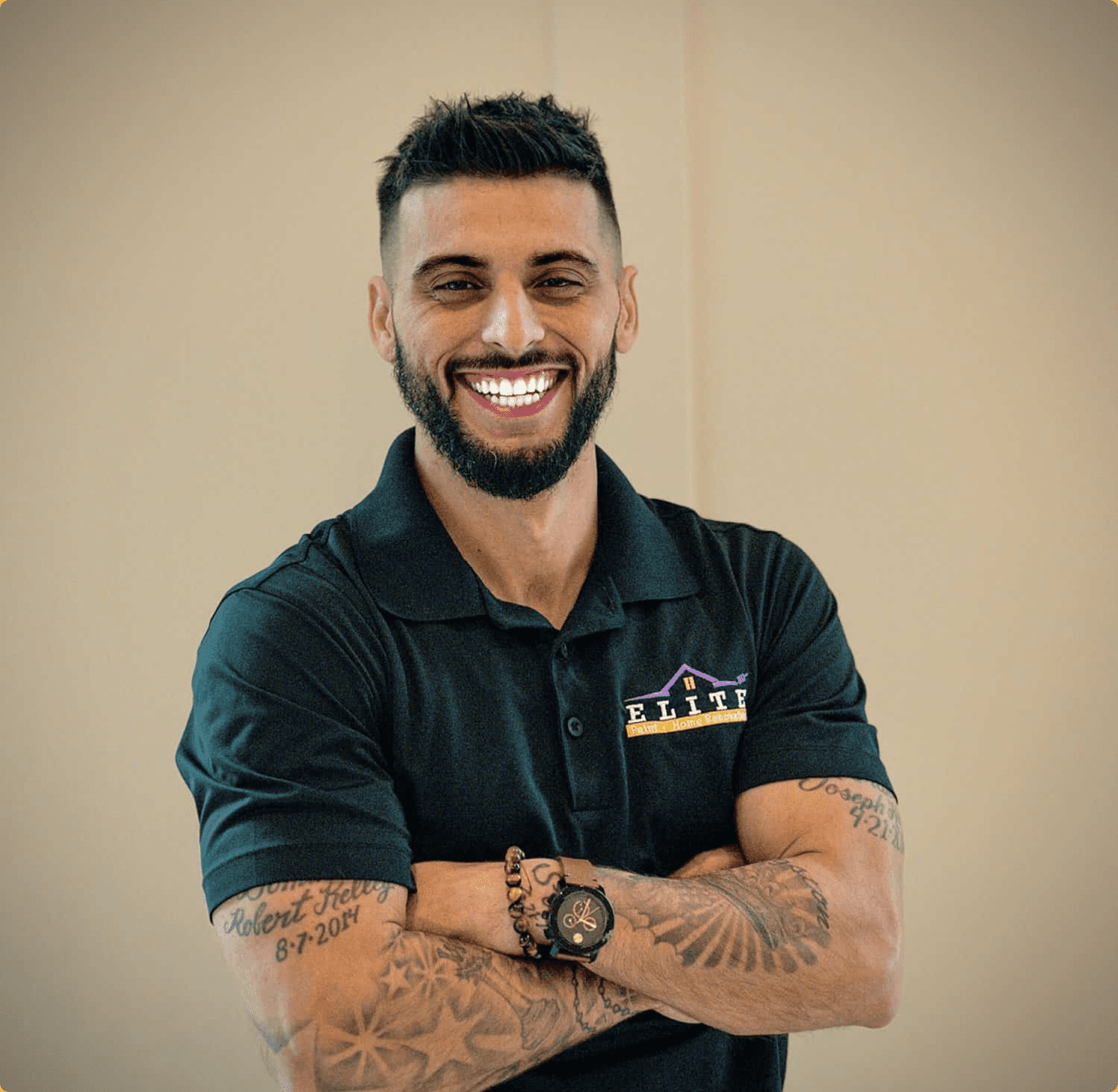 Man with tattoos smiling, wearing a logoed polo shirt, arms crossed, well-lit setting.
