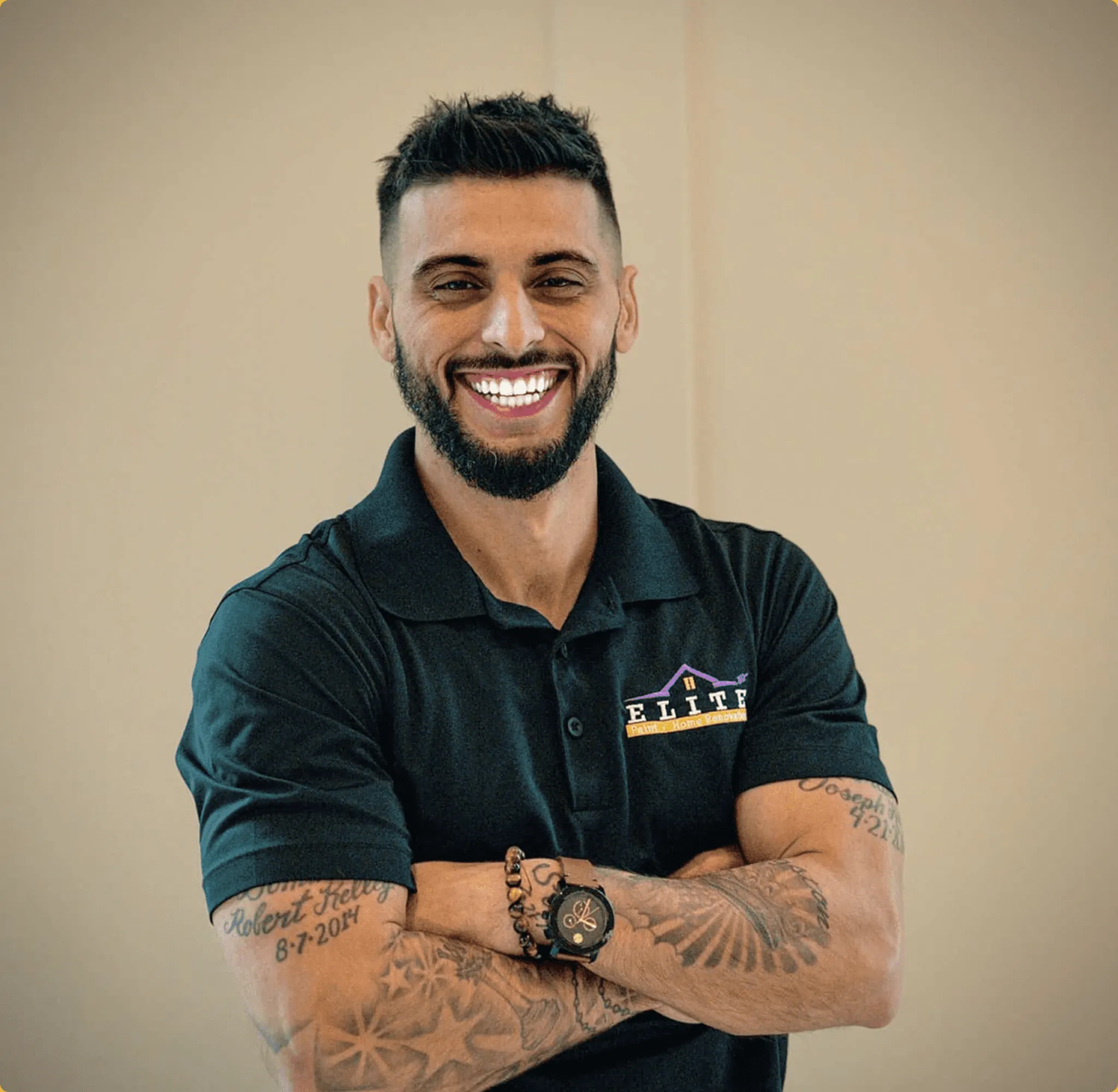 Man with tattoos smiling, wearing a logoed polo shirt, arms crossed, well-lit setting.